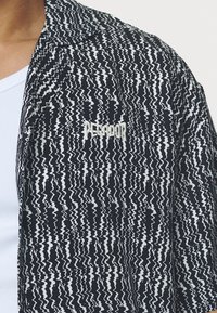 Svartvit jacka med zigzag-mönster, tillverkad av tyg med en texturerad yta, med en subtil logotyp broderad på bröstet.