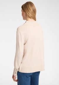 Vaaleanbeige neule, jossa on ribbikaulus, pitkät hihat ja koristeelliset napit hihojen päällä. Pehmeä, sileä pinta ja rento istuvuus.