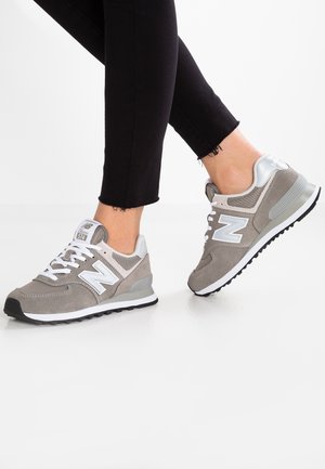 Person iført grå og hvide New Balance sneakers med sorte bukser står på en hvid overflade.