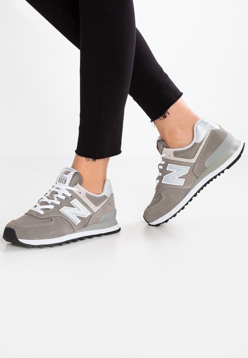 Personne portant des baskets New Balance grises et blanches avec un pantalon noir, debout sur une surface blanche.