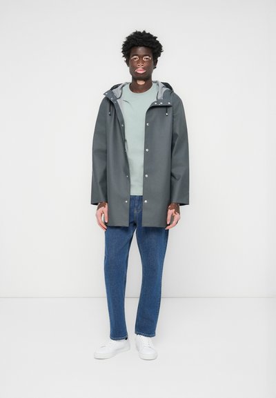 Grå vanntett jakke med hette, sølvknapper, lys grå fôr, båret over en lys grønn skjorte, kombinert med blå jeans og hvite sneakers.