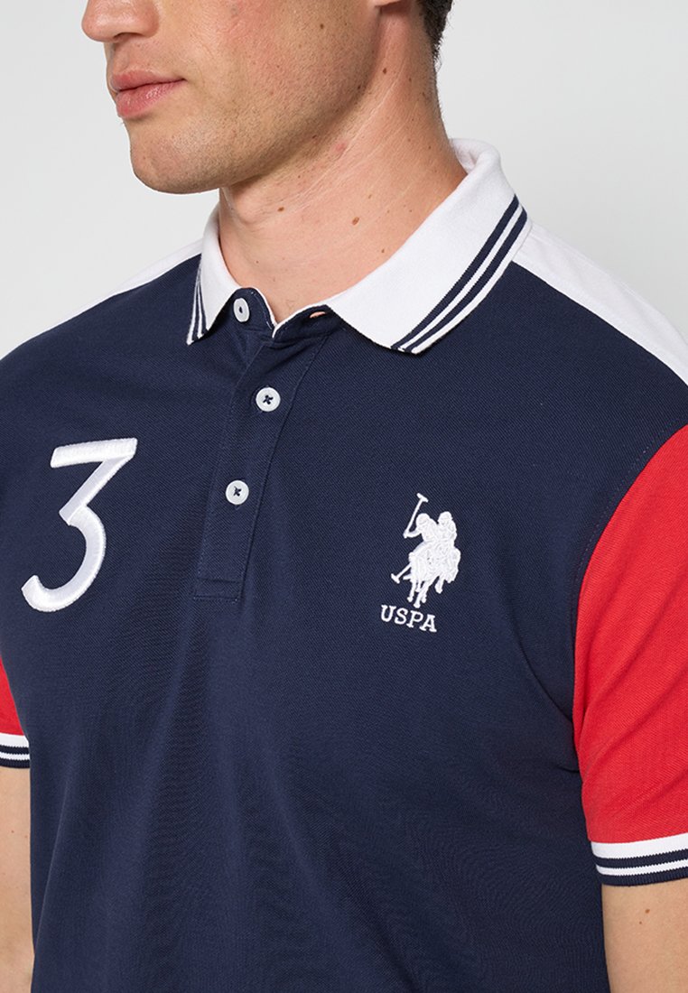 Polo-shirt marine avec des manches rouges et un col blanc. Présente le numéro brodé "3" et le logo USPA. Matière en coton doux, coupe classique.