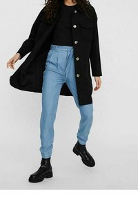 Abrigo largo negro con botones plateados, combinado con pantalones ajustados azul claro y botas de tobillo negras. El conjunto presenta un diseño simple y estructurado.