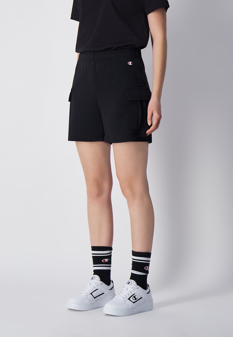 Shorts atletici neri in cotone con vita regolabile, tasche laterali e vestibilità comoda, abbinati a sneaker bianche e calze a righe.