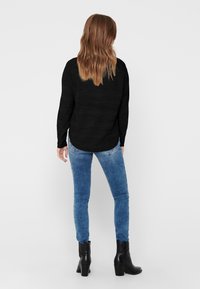 Schwarzes Langarmoberteil mit strukturiertem Design, kombiniert mit hellblauen Skinny-Jeans und schwarzen, knöchelhohen Stiefeln mit Absatz, von hinten betrachtet.