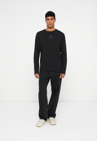 Melnā garajaka ar apaļu kakla izgriezumu, uz kuras krūšu daļas ir zila rakstība, apvienota ar tumšām džinsām un baltām kedām.