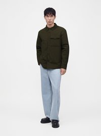 Jeune homme debout portant une chemise à boutons vert foncé avec une poche poitrine, un jean large bleu clair et des chaussures noires épaisses.