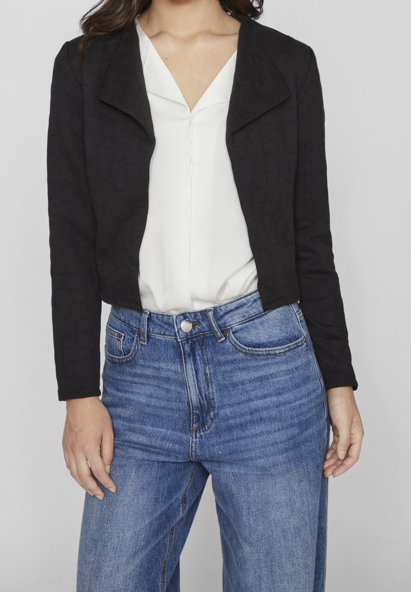 Femme portant une veste courte noire par-dessus un chemisier blanc et un jean bleu taille haute, debout devant un fond uni gris clair.