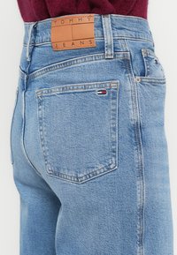 Világoskék egyenes szabású farmernadrág. Barna bőrcímkével rendelkezik, amelyen a "TOMMY JEANS" felirat olvasható, valamint egy kis zászlólogó található a hátsó zsebnél.