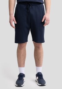 Shorts navy en tissu doux, dotés d'une taille élastique avec cordon de serrage, de poches avant et d'une coupe jusqu'aux genoux, associés à des chaussures de sport.