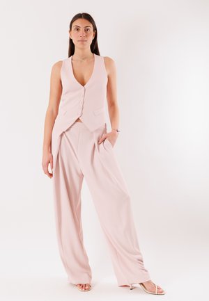 Donna in piedi con la mano in tasca, indossa un gilet rosa pallido senza maniche con bottoni, pantaloni a gamba larga e sandali con tacco beige su sfondo bianco.