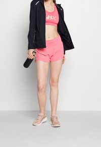 Sportoutfit mit einer schwarzen Jacke, einem rosa Sport-BH und rosa Shorts. Kombiniert mit rosa Laufschuhen und einem schwarzen Wasserflasche.