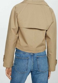 Giacca cropped beige con dettagli di pattina anti-tempesta indossata con jeans in denim blu a vita alta, mostrata da dietro.