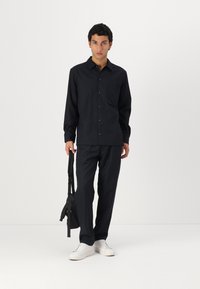Dark navy button-up shirt with a chest pocket, paired with matching trousers. Features a modern fit and worn with white sneakers and a black bag.