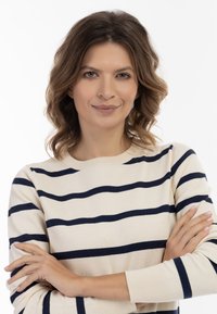 Beżowy sweter w granatowe paski, okrągły dekolt i długie rękawy. Gładka tekstura z prążkowanymi mankietami. Modelka pozuje z założonymi rękami.