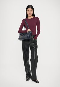 Top de culoare burgundy cu mâneci lungi, pantaloni negri cu talie înaltă din piele ecologică și balerini negri. Modelul ține în mână o poșetă din piele albastră închisă cu un breloc.