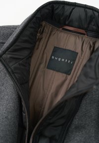 Graue Außenstoffjacke mit einem schwarzen, gequilten Innenfutter. Verfügt über ein Etikett mit "bugatti" in weißem Text. Glatte Textur und taillierte Form.