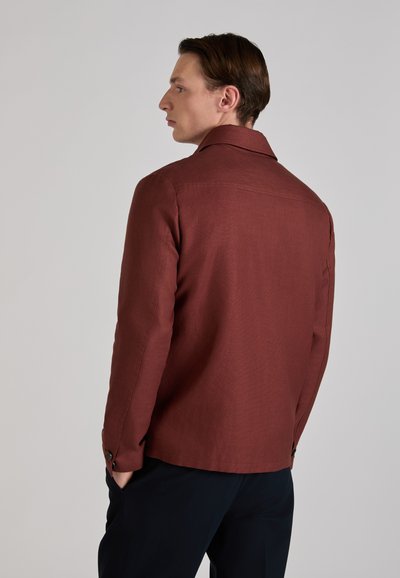 Mężczyzna w brązowej kurtce i czarnych spodniach, z rękami w kieszeniach, stojący bokiem i patrzący w lewo na jednolitym szarym tle.