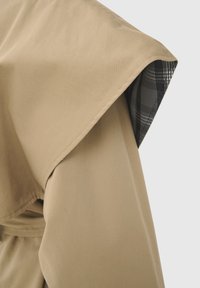 Detaliu pe umărul unui trench bej care arată clapeta pliată și căptușeala interioară în carouri alb-negru.