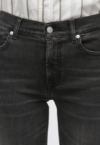 Fekete farmernadrág, karcsúsított szabással, gombos záródással, övbújtatókkal és első zsebekkel. A szövet enyhén texturált megjelenésű.