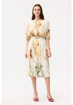 Une femme se tient debout, portant une robe midi crème à imprimé floral jaune, manches courtes bouffantes, ceinture beige, et des sandales à talons ouvertes rouges.