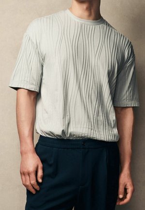 Helles graues T-Shirt mit gewelltem Texturmuster, Rundhalsausschnitt und kurzen Ärmeln, kombiniert mit dunkelblauen Hosen und einem Knopfdetails.