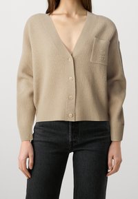 Beige ribbstickad kofta med djup V-ringning, knappar fram och en ficka. Har långa ärmar och mjuk textur.