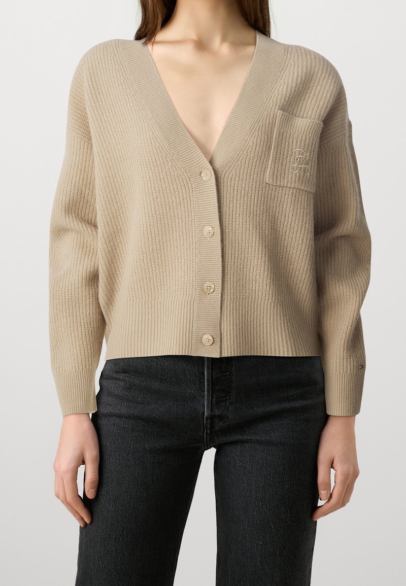 Beige ribbstickad kofta med djup V-ringning, knappar fram och en ficka. Har långa ärmar och mjuk textur.