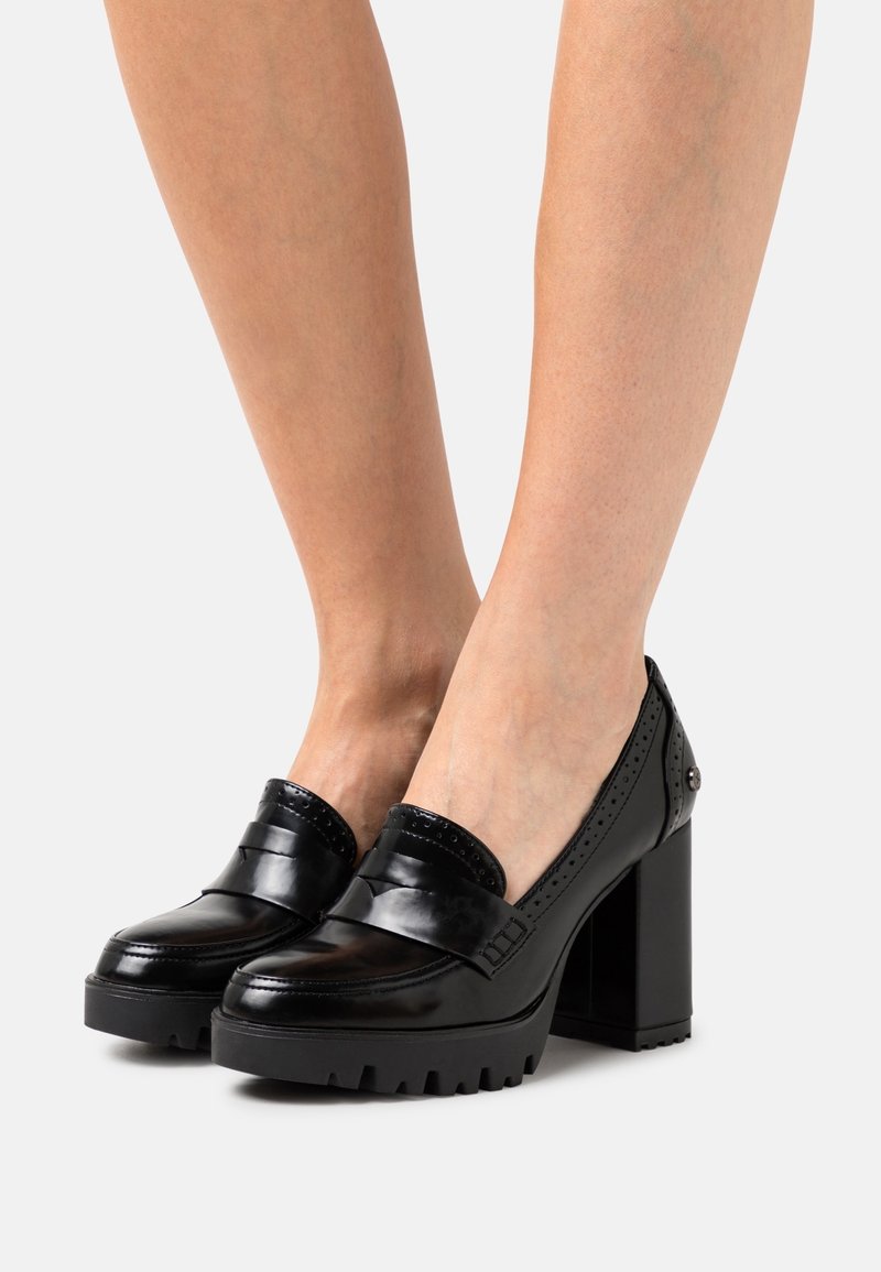 Chaussures à talons en cuir verni noir avec un talon carré chunky, une semelle à grip texturé et des perforations décoratives sur le dessus.