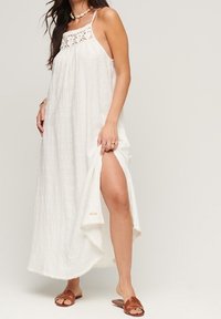 Vestido maxi blanco ligero con parte superior de ganchillo y tirantes finos. Presenta una abertura lateral y un dobladillo con bordes festoneados. Combinado con sandalias marrones.