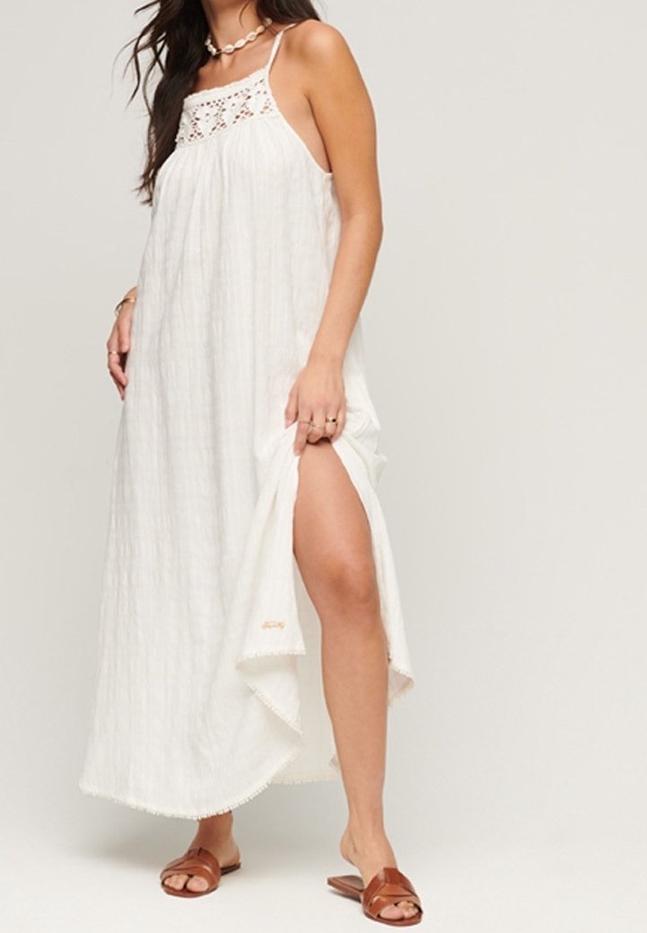 Vestido maxi blanco ligero con parte superior de ganchillo y tirantes finos. Presenta una abertura lateral y un dobladillo con bordes festoneados. Combinado con sandalias marrones.