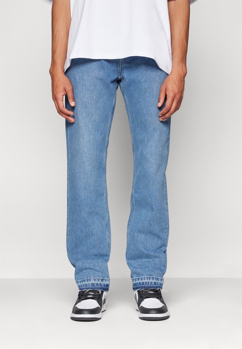 Los jeans de mezclilla azul claro presentan un diseño de pierna recta, cinco bolsillos y un acabado desgastado. Combinados con zapatillas negras y blancas.