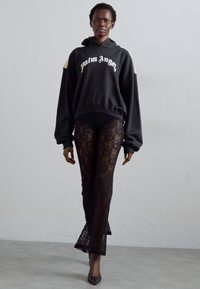 Black hoodie with "Palm Angels" logo, lace leggings featuring a floral pattern, and pointed heels. Model stands against a neutral background.