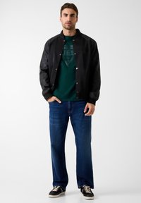 Czarna kurtka bomberka z teksturowanymi rękawami, zielona koszulka z logo, niebieskie jeansy o prostych nogawkach oraz czarne trampki z kremowymi akcentami.