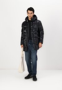 Jachetă puffer neagră cu un finisaj lucios, glugă și închidere cu nasturi, purtată cu blugi denim închis și pantofi negri, ținând o geantă din pânză.