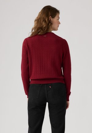 Femme aux cheveux ondulés portant un pull rouge texturé et un jean noir, debout de dos devant un fond clair uni.