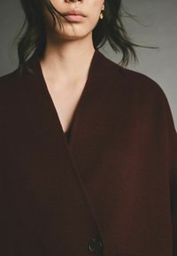 Burgundy gyapjú kabát mély V nyakkivágással, egyetlen fekete gombbal és puha, texturált felülettel.