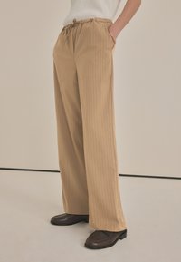 Beige gestreifte weite Hosen mit elastischem Bund und Seitentaschen, kombiniert mit braunen Schuhen, vor einem neutralen Hintergrund.