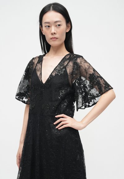 Juoda nėriniuota suknelė su giliu V formos iškirpimu ir plazdančiomis rankovėmis. Papuošta gėlių raštais ir subtiliais blizgučiais, demonstruojanti tekantį siluetą.