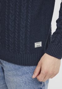 Pull en mailles côtelées bleu marine avec un ourlet et des poignets à côtes, porté avec un jean bleu clair ; petite étiquette blanche sur le bas avant indique "11 Project."