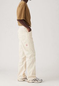 Pantaloni cargo beige chiaro con tasche laterali, abbinati a una camicia marrone a maniche corte e sneakers grigie con upper in rete e dettagli a fantasia.