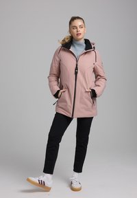 Roze geïsoleerde jas met een zwarte capuchon. Heeft een voorkeursluiting met rits, twee zijzakken en is gecombineerd met zwarte broek en witte sneakers.