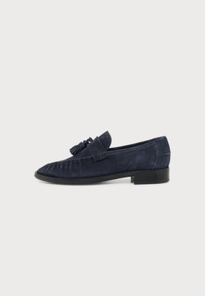 Mocassin en daim bleu marine avec détail pompon, talon bas noir et dessus cousu style mocassin, présenté en profil latéral sur fond blanc.