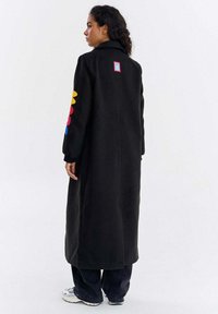 Kobieta stojąca bokiem, ubrana w długi czarny płaszcz z kolorowymi kwiatowymi łatami na lewym rękawie oraz małą łatą na górnej części pleców.