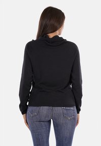 Maglione nero lavorato a maglia con collo a scialle, forma aderente, maniche lunghe e texture morbida. Indossato sopra jeans blu, in vista posteriore.