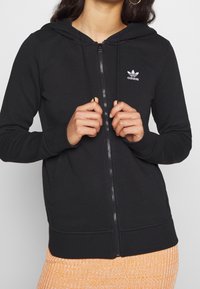 Svart zip-up hoodie i mjukt tyg med dragkedja fram, huva med snörning och en vit Adidas-logotyp på övre vänstra sidan.