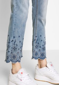 Jean en denim bleu clair avec des ourlets festonnés, ornés de délicates broderies florales bleu foncé et d'une bordure effilochée. Associé à des baskets blanches.