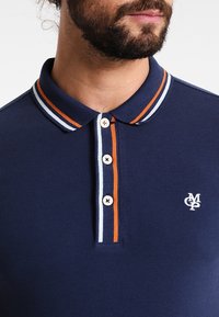 Námořnické polo tričko s kontrastním oranžovým a bílým proužkovaným límcem, zapínáním na tři knoflíky a vyšitým logem na levé straně hrudi.