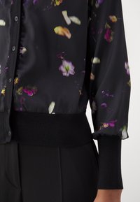 Blouse noire transparente à fleurs avec manches longues bouffantes, devant boutonné, poignets et ourlet côtelés, portée avec un pantalon noir.