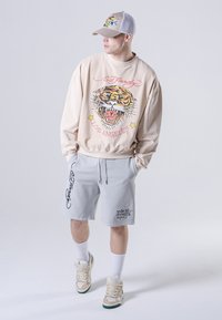Uomo che indossa una felpa beige Ed Hardy con grafica di una tigre, pantaloni corti grigi, scarpe da ginnastica beige, calzini bianchi e un cappellino beige e bianco, mentre guarda di lato.